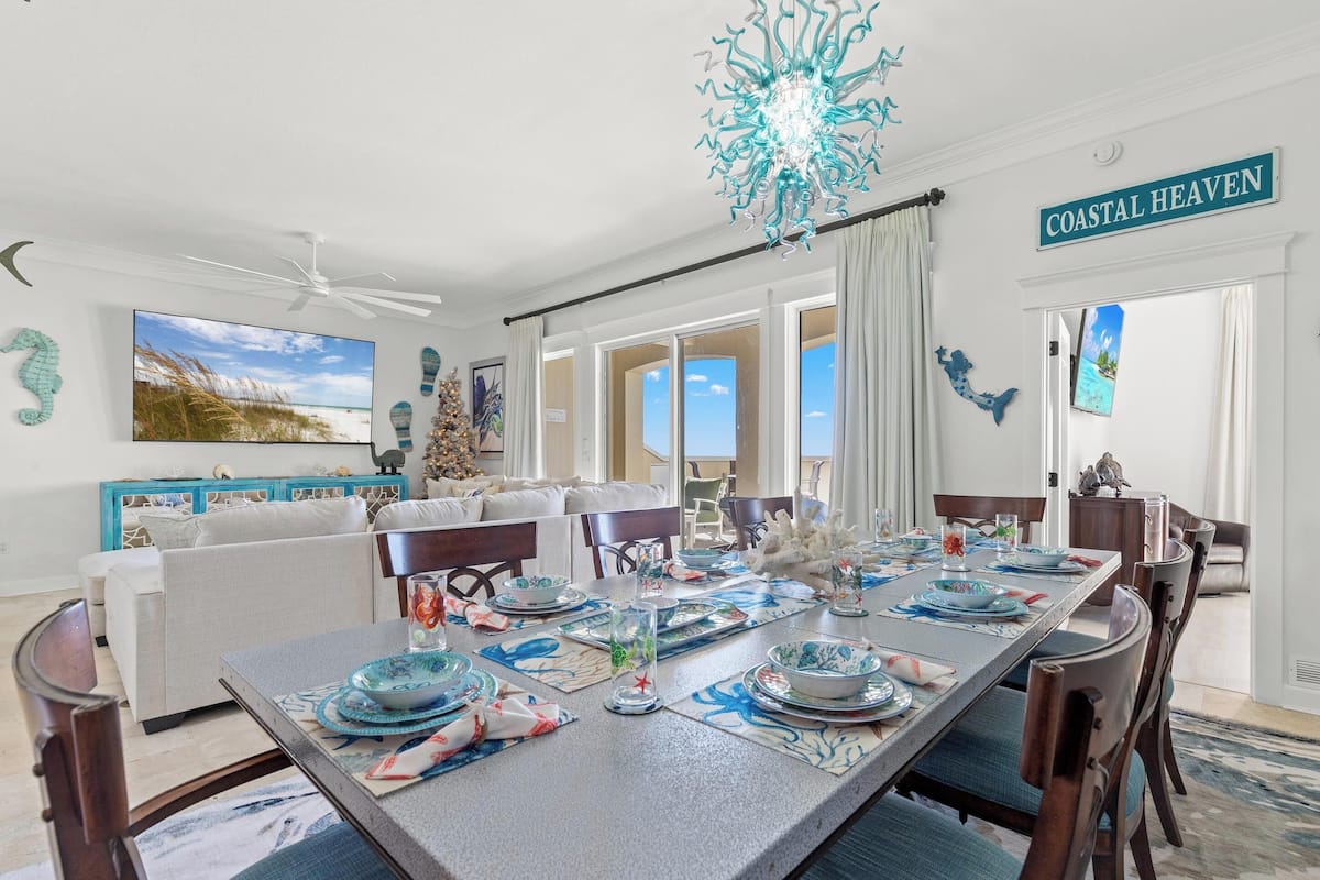 A dining area with a large table set for six, surrounded by chairs. There is a ceiling fan, a chandelier, and a sign that reads "COASTAL HEAVEN".
