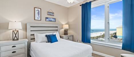 A bedroom with a bed, bedside tables, a nightstand, a chair, a window with a view of the beach, and a ceiling fan.