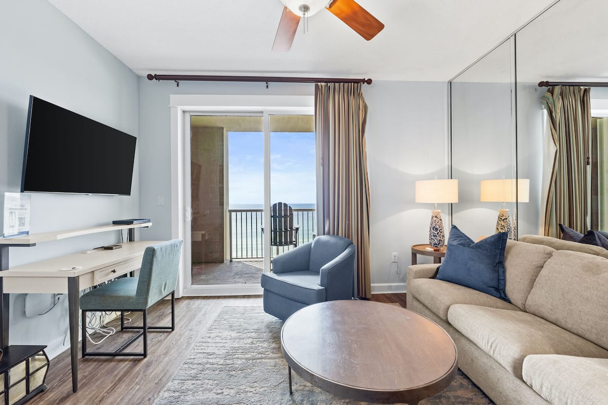 A hotel room with a flat-screen TV, a desk with a chair, a sofa, a round coffee table, a ceiling fan, and a balcony with curtains.