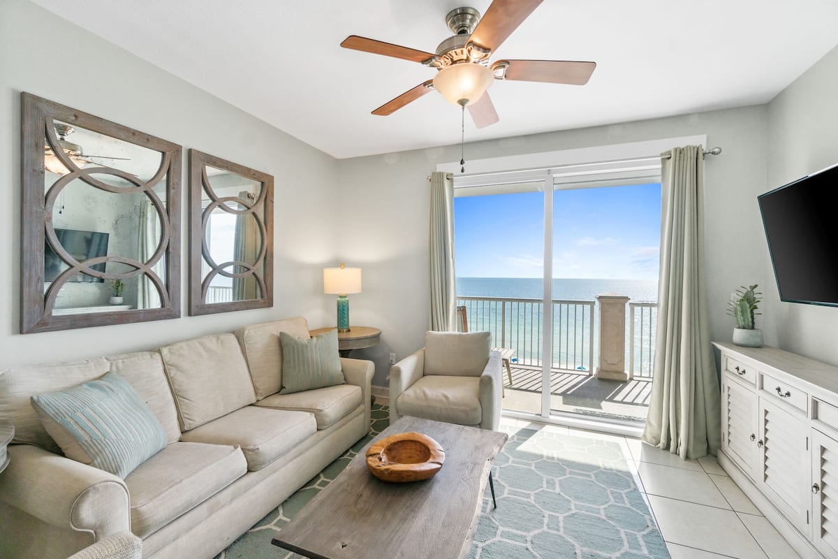 A room with a ceiling fan, a sofa, a coffee table, a TV, and a view of the ocean through a sliding glass door.