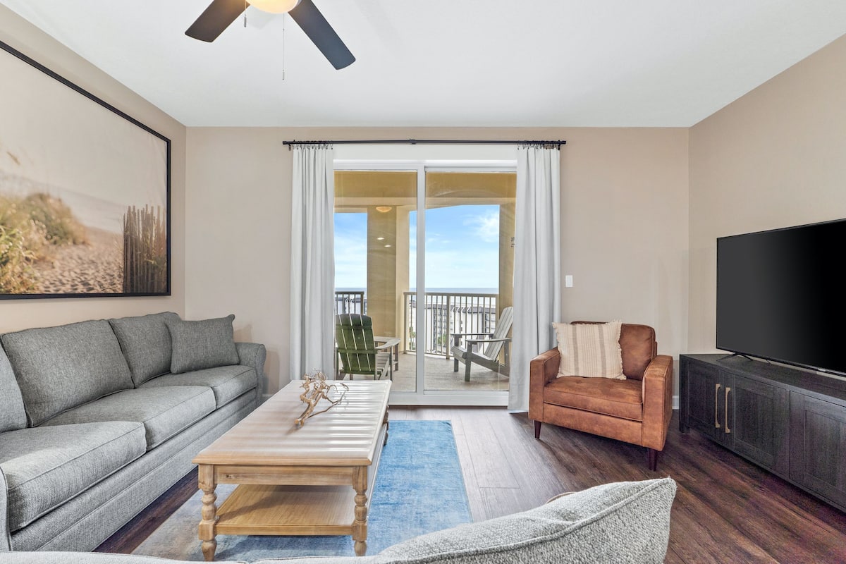 A living room with a gray sofa, a wooden coffee table, a brown armchair, a flat-screen TV, and a balcony with a view.