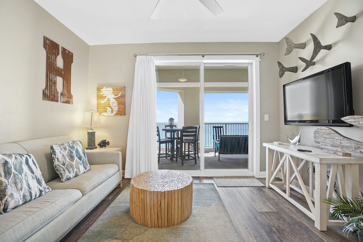 A living room with a sofa, a round wooden ottoman, a TV, and a balcony with a table and chairs.