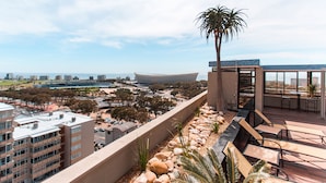 A rooftop terrace with a palm tree, seating area, and a view of a cityscape including a stadium. - Unit Number 318 Cape Royale Luxury Apartments (Cape Town)