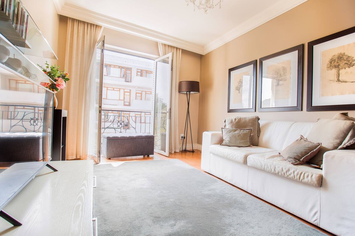Een moderne woonkamer met een witte bank, een glazen salontafel en een vloerlamp. Er is een balkon met uitzicht op gebouwen en een deur naar buiten.