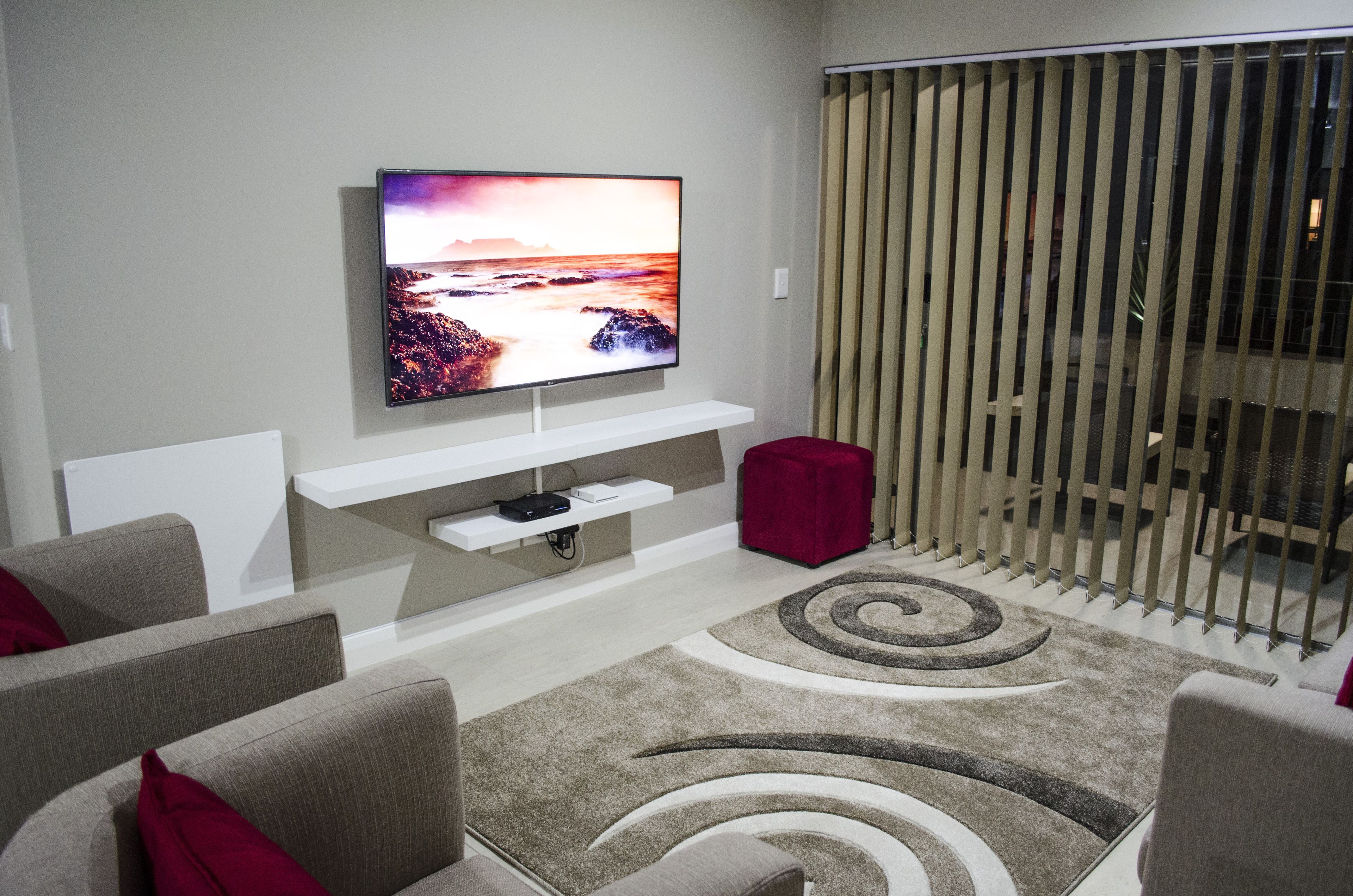 A modern living room with a flat-screen TV mounted on a white shelf, grey sofas with red cushions, a patterned rug, and a sliding glass door with vertical blinds.