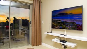 En moderne stue med et stort fladskærms-tv monteret på en hvid reol, en skydedør i glas med et brunt gardin, og en lille hvid telefon på en hylde under tv'et.