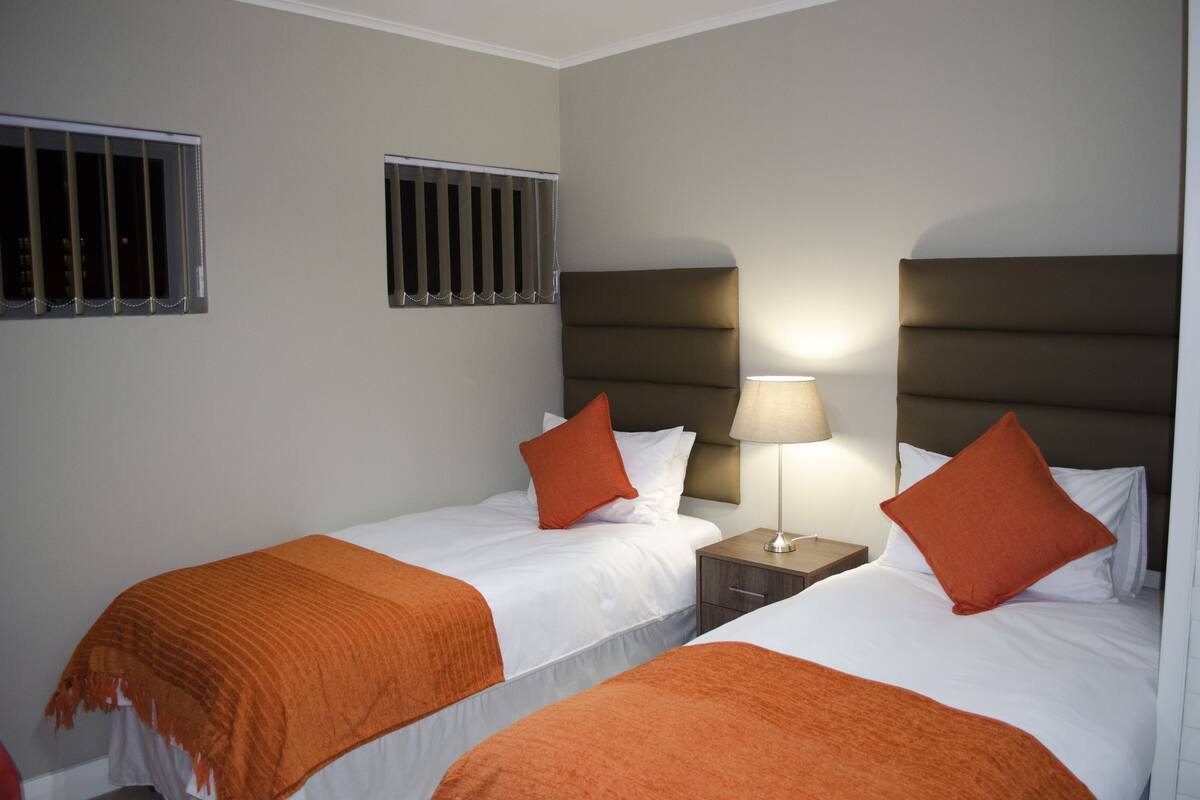 A hotel room with two beds, each with an orange blanket and pillow, a nightstand with a lamp, and barred windows.