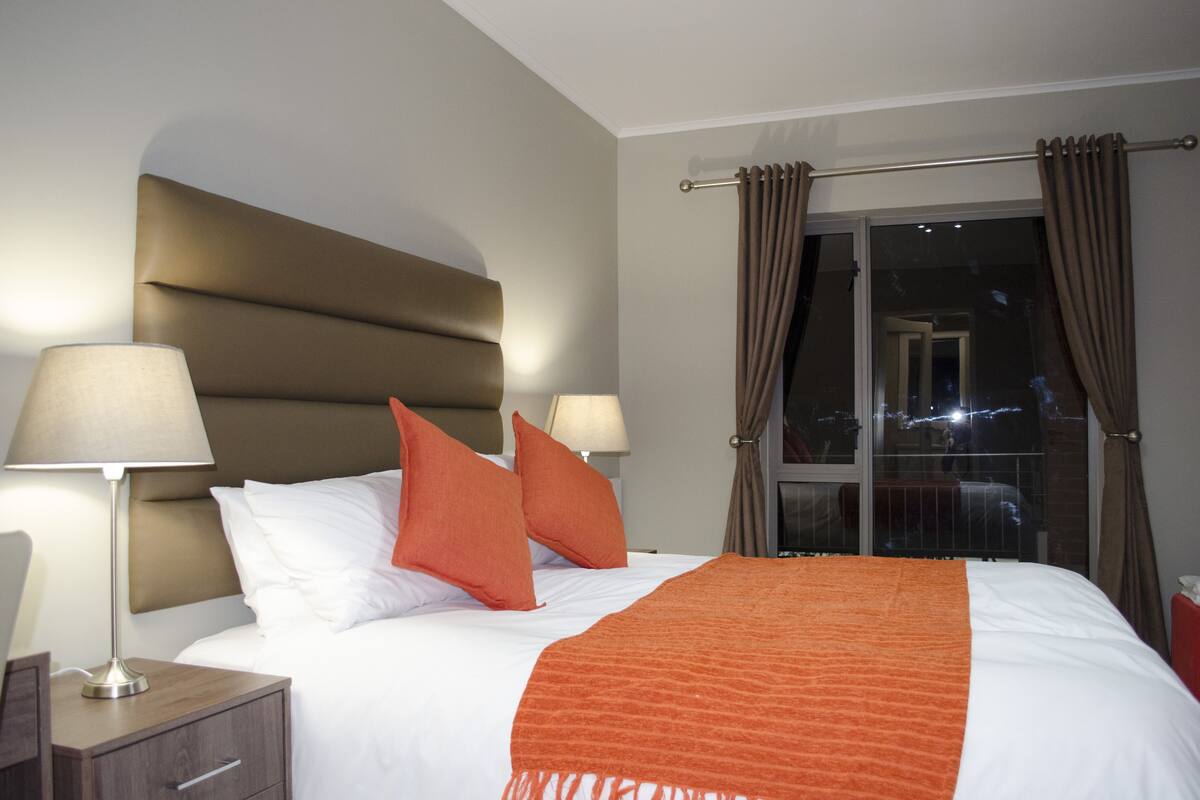 A hotel room with a bed, two bedside lamps, a window with curtains, and a nightstand with a lamp.