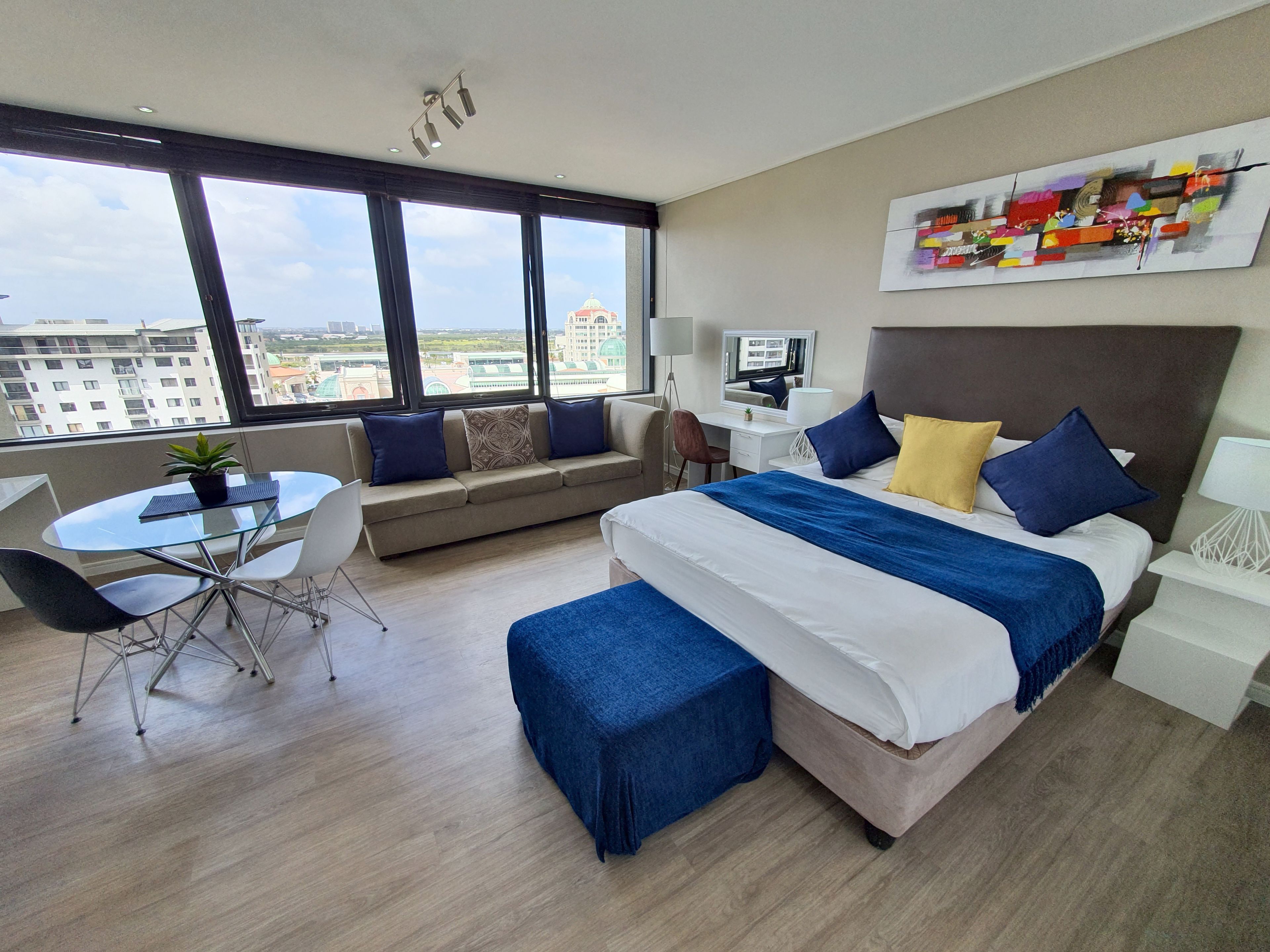Een moderne hotelkamer met een groot bed, een slaapbank, een eettafel en uitzicht op de stad door grote ramen.