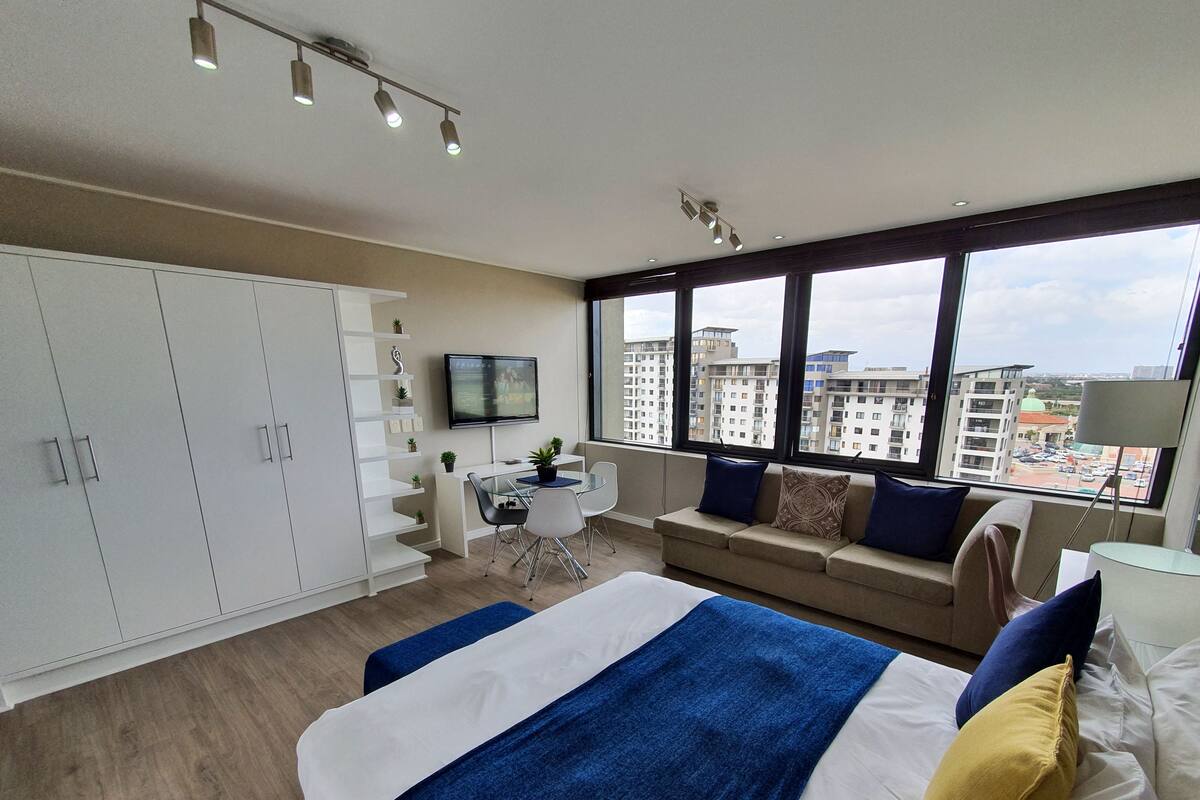 A modern hotel room with a large bed, a desk, and a sofa. There are multiple windows offering a city view.
