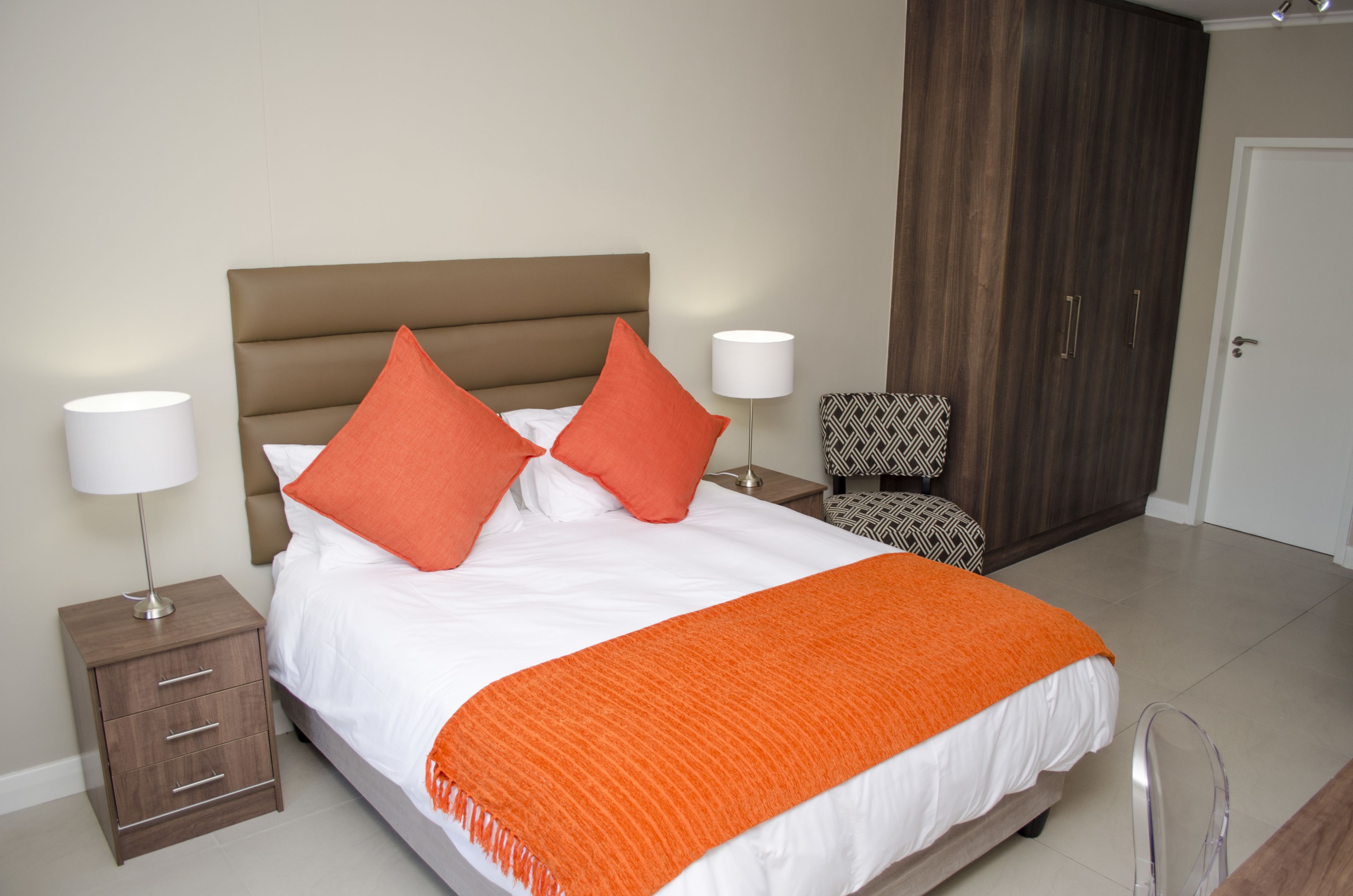 A neatly made bed with two pillows, a bedside table with a lamp, a patterned chair, and a wardrobe.