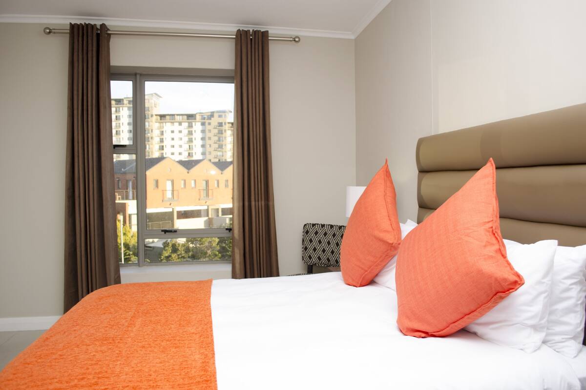 A hotel room with a large bed, orange and white bedding, a view of buildings through a window with brown curtains, and a headboard with a beige upholstered panel.