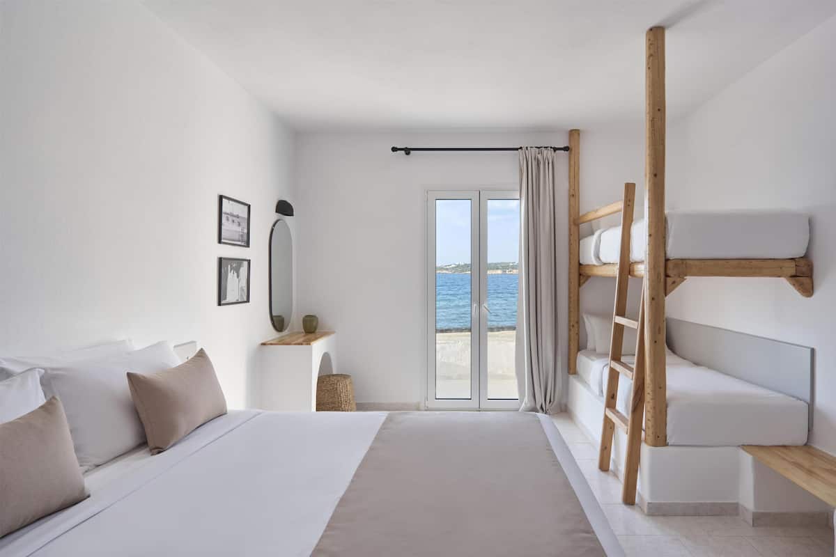 A bedroom with a bed, a wooden ladder, a small table, and a view of the sea through a sliding door.
