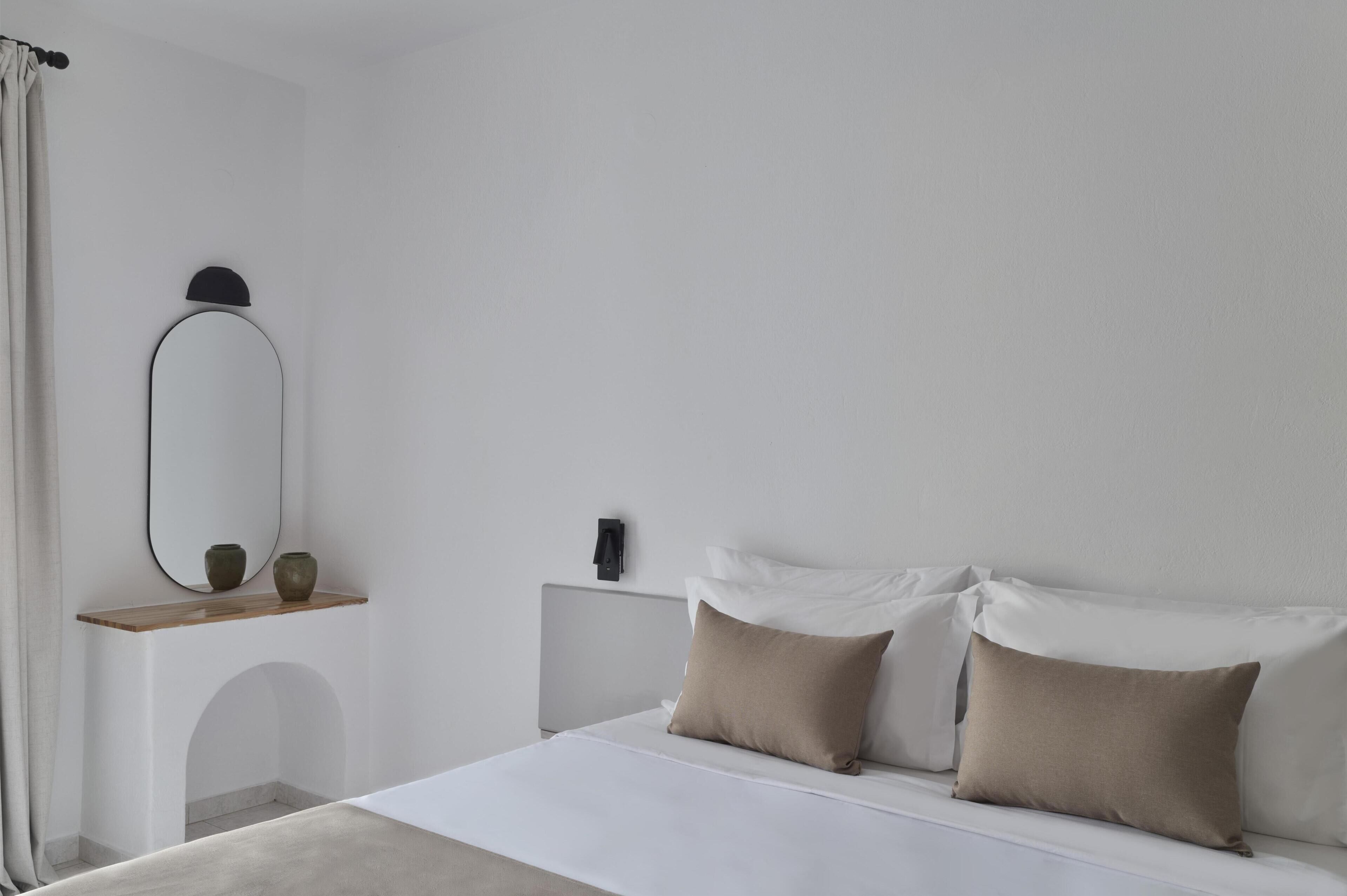 A minimalist bedroom with a bed, two pillows, a nightstand, a mirror, and a shelf with two vases.