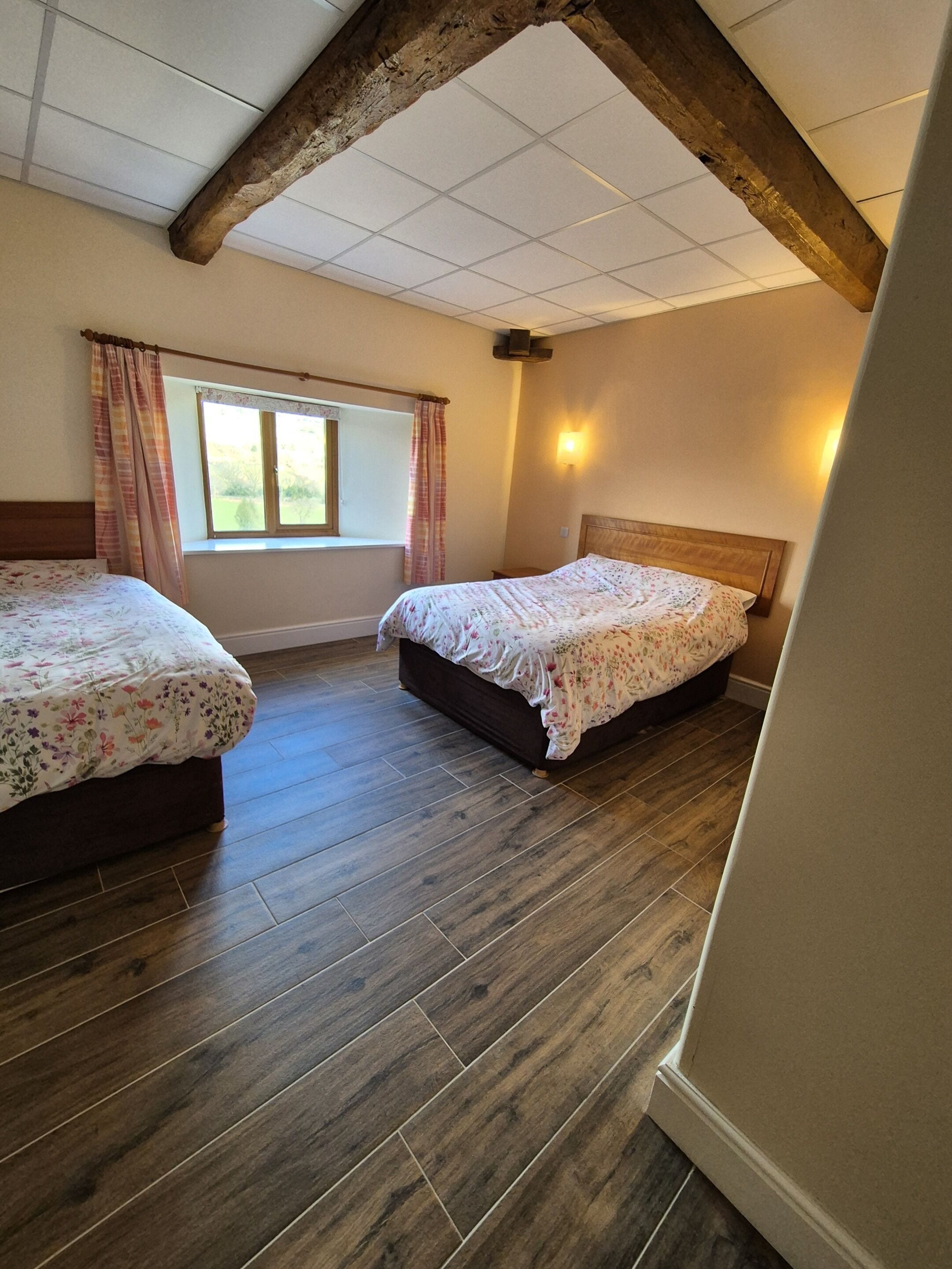 Ein Schlafzimmer mit zwei Betten, Holzboden, einem Fenster mit Vorhängen und einer schrägen Decke mit freiliegenden Balken.
