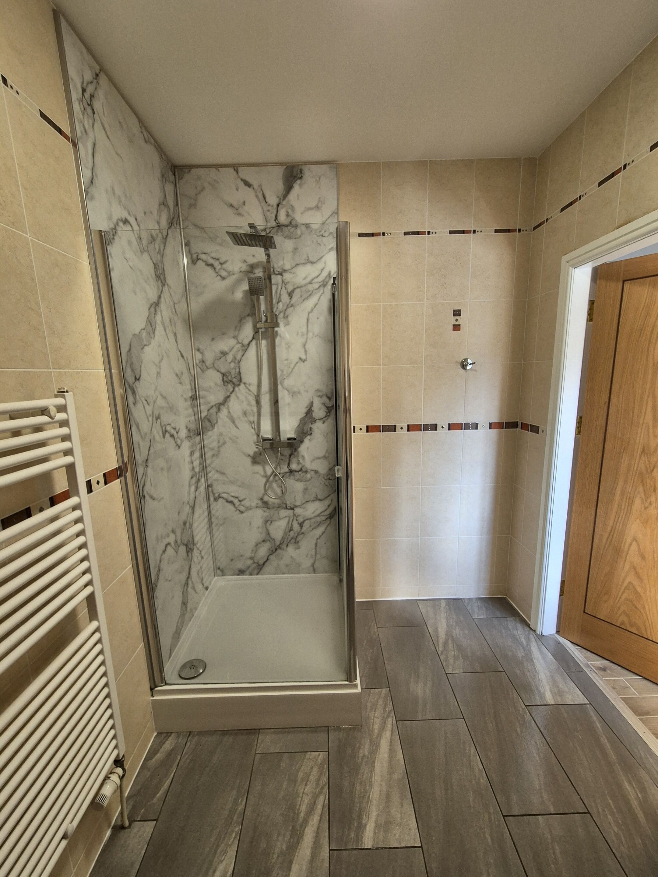 A shower with a glass door, a white radiator, and a wooden door in a bathroom with tiled walls and floors.