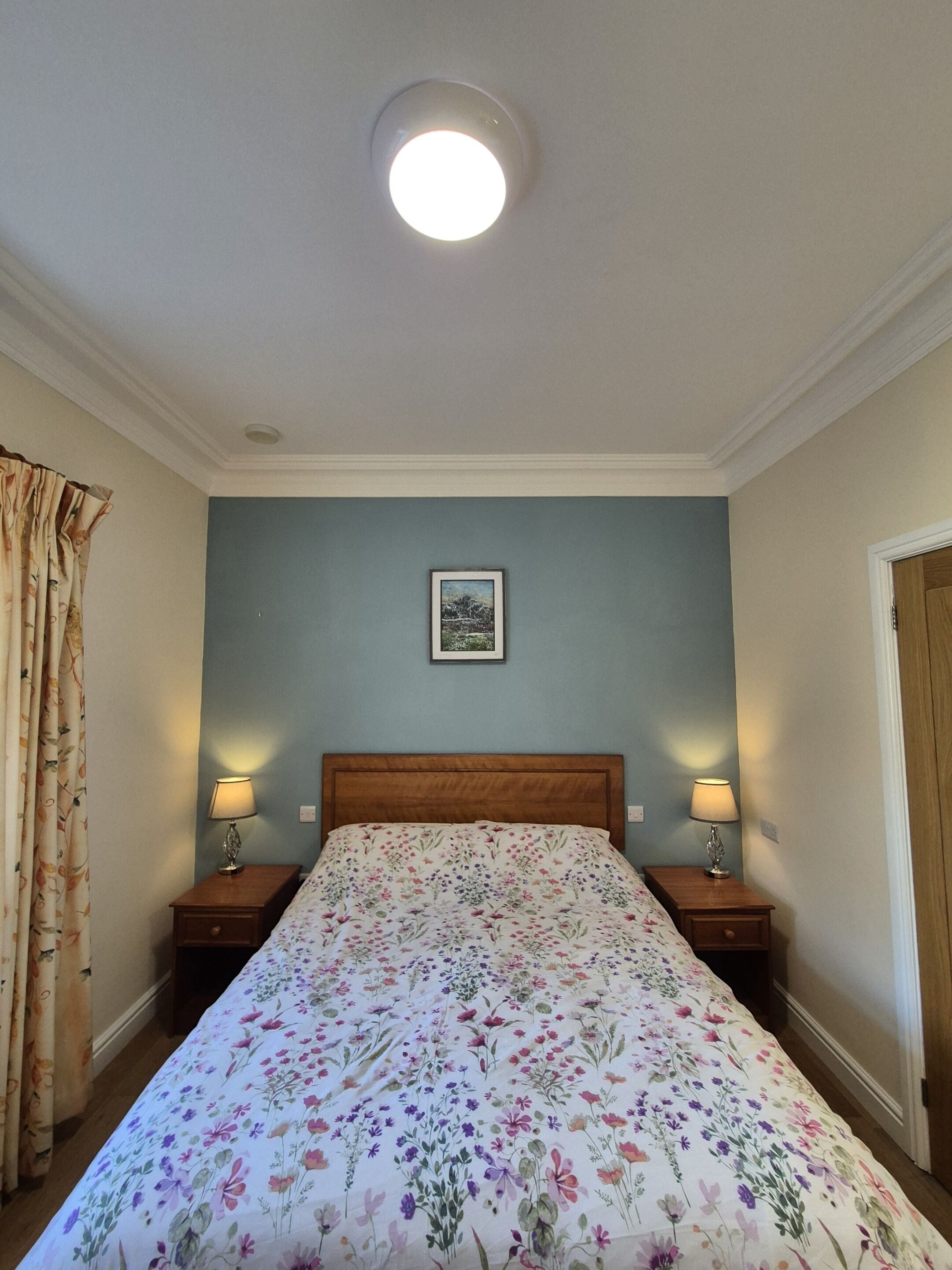 A bedroom with a floral bedspread, a wooden headboard, two bedside lamps, a framed picture on the wall, and a ceiling light.