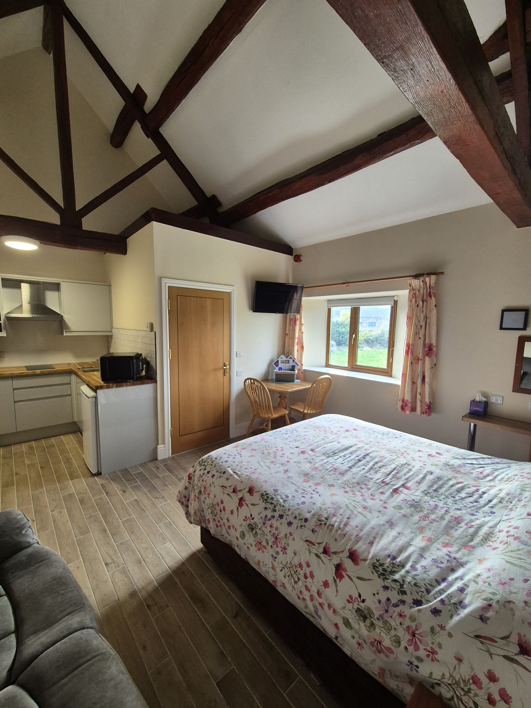A compact bedroom with a bed, a small desk, a chair, a window with curtains, and a kitchenette area.