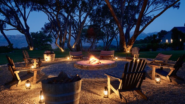 A fire pit surrounded by Adirondack chairs and lanterns, set in a gravel area with trees and a mountain view in the background.