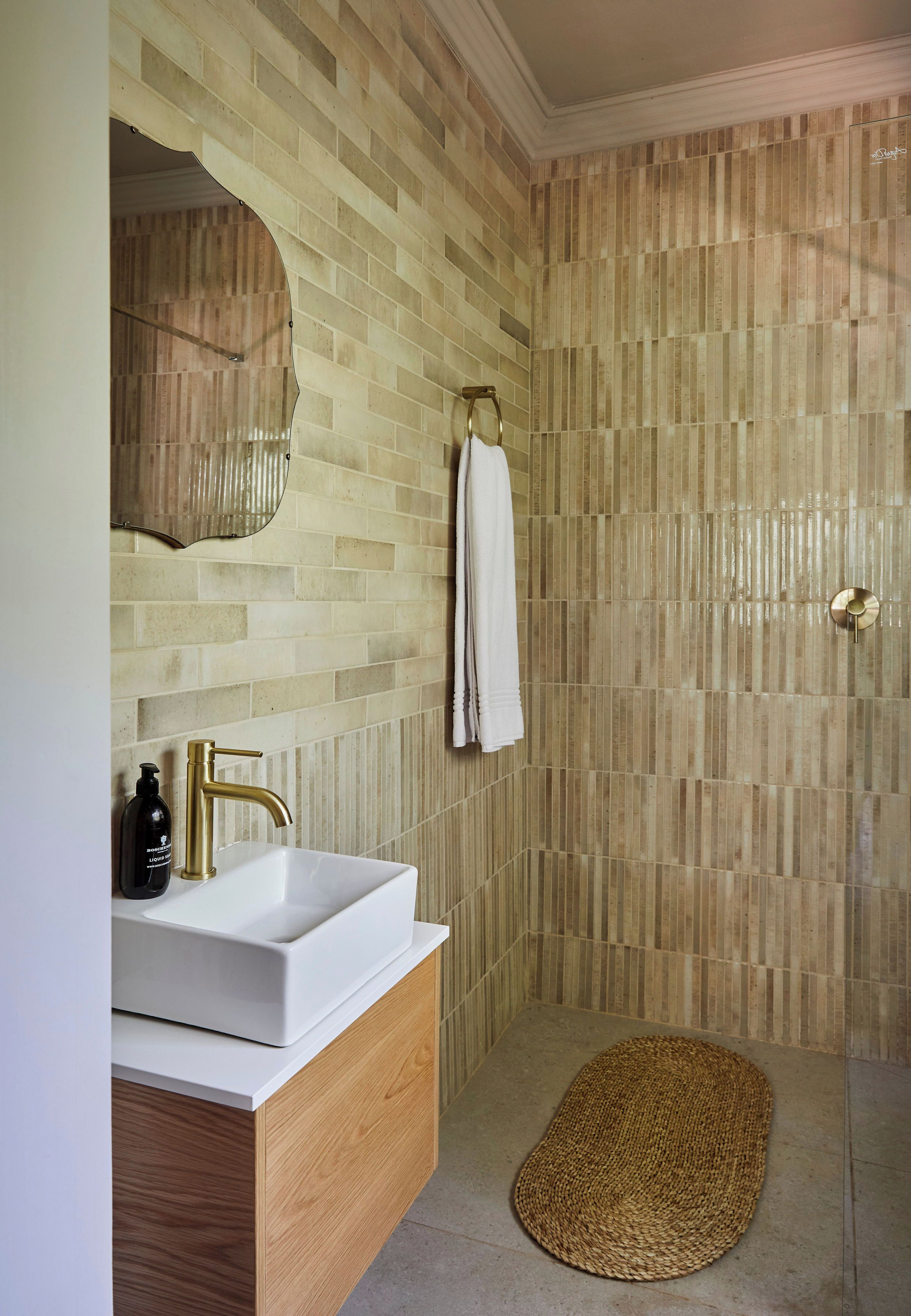 Un baño con lavamanos blanco, grifo dorado y tocador de madera. Una toalla cuelga de un perchero, y un felpudo está en el suelo.