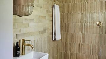 Un baño con lavamanos blanco, grifo dorado y tocador de madera. Una toalla cuelga de un perchero, y un felpudo está en el suelo.