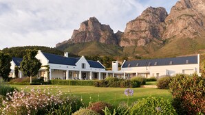 A white building with a dark roof, surrounded by a well-maintained garden and mountains in the background.
