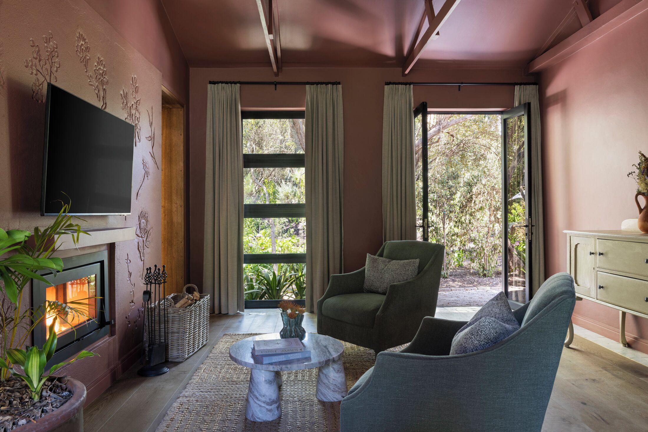 A cozy living room with a fireplace, comfortable seating, and large windows offering a view of greenery.