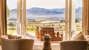 A room with a large sliding glass door leading to a scenic outdoor view of mountains, a grassy field, and a lake. Inside, there is a wicker sofa with cushions and a small table with a book and a vase.