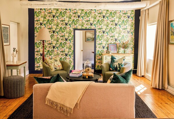 A living room with floral wallpaper, two green armchairs, a beige sofa, a small round ottoman, and a side table with a lamp. - Boschendal Farm Estate (Franschhoek)
