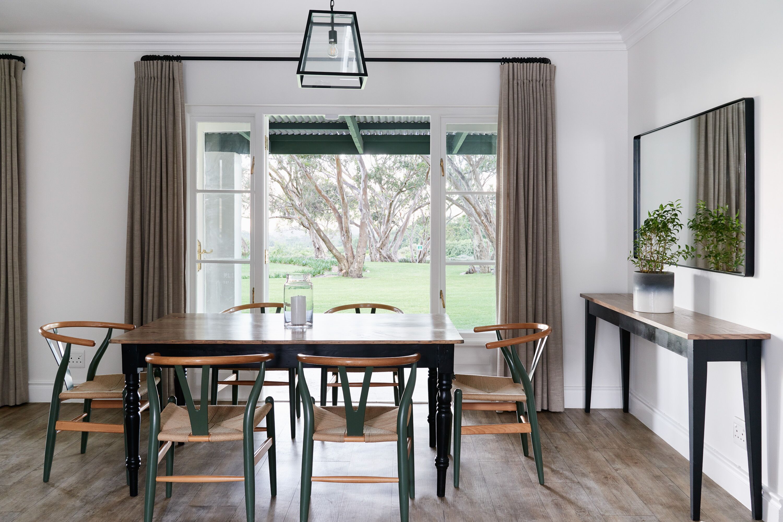 A dining area with a wooden table and chairs, a hanging light fixture, and a view of a garden through a window.