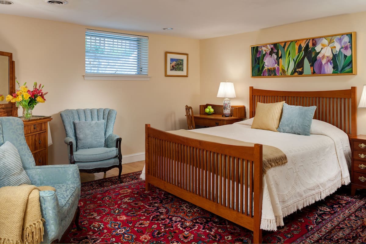 A bedroom with a wooden bed, a blue armchair, a wooden nightstand, a vase with flowers, a framed picture, and a large painting on the wall.