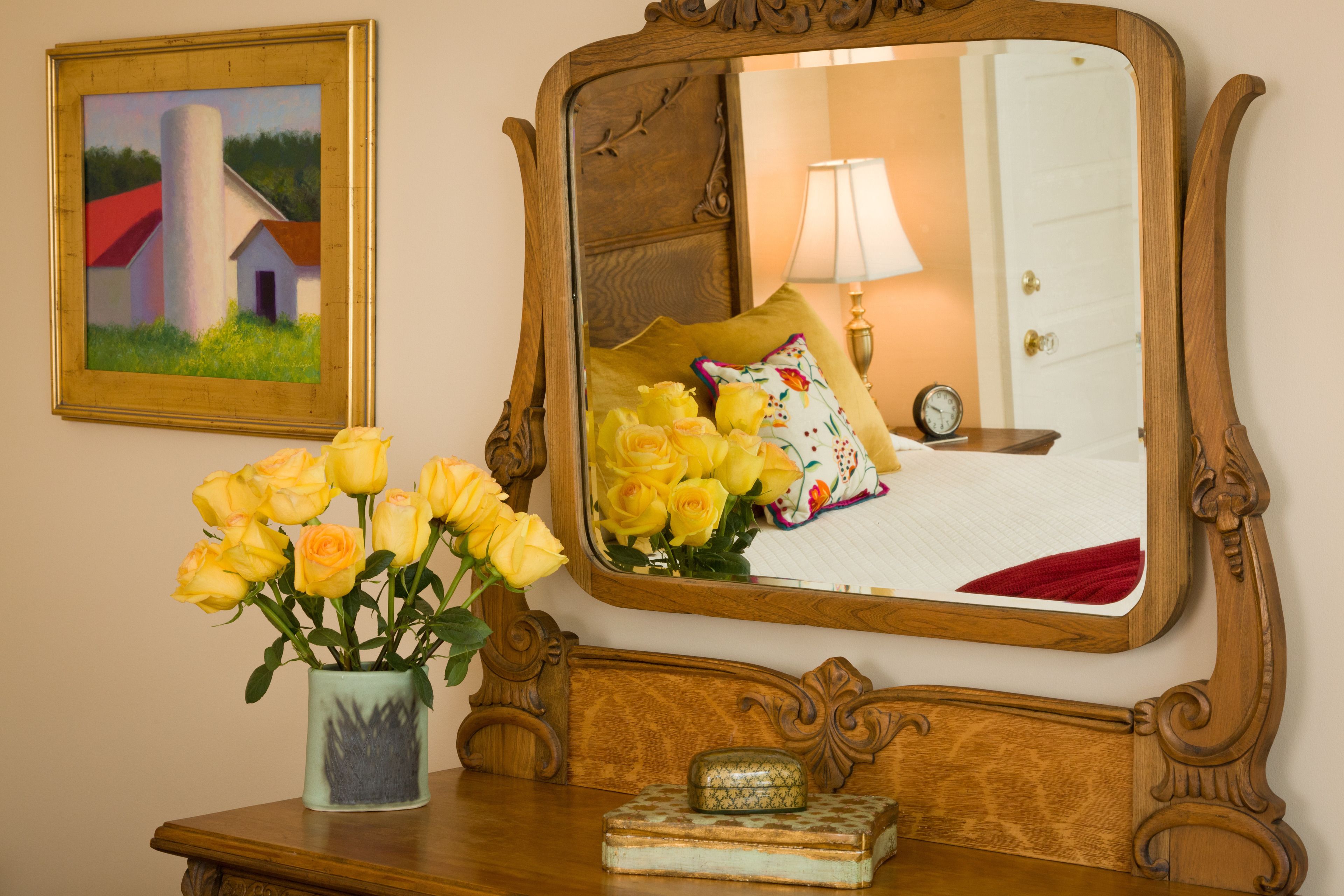 A wooden bedside table with a vase of yellow roses, a framed painting, and a mirror reflecting a room with a bed, lamp, and clock.