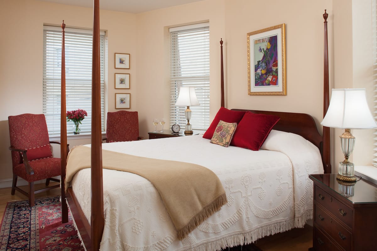 A bedroom with a four-poster bed, a red armchair, a nightstand with a lamp, and a window with blinds.