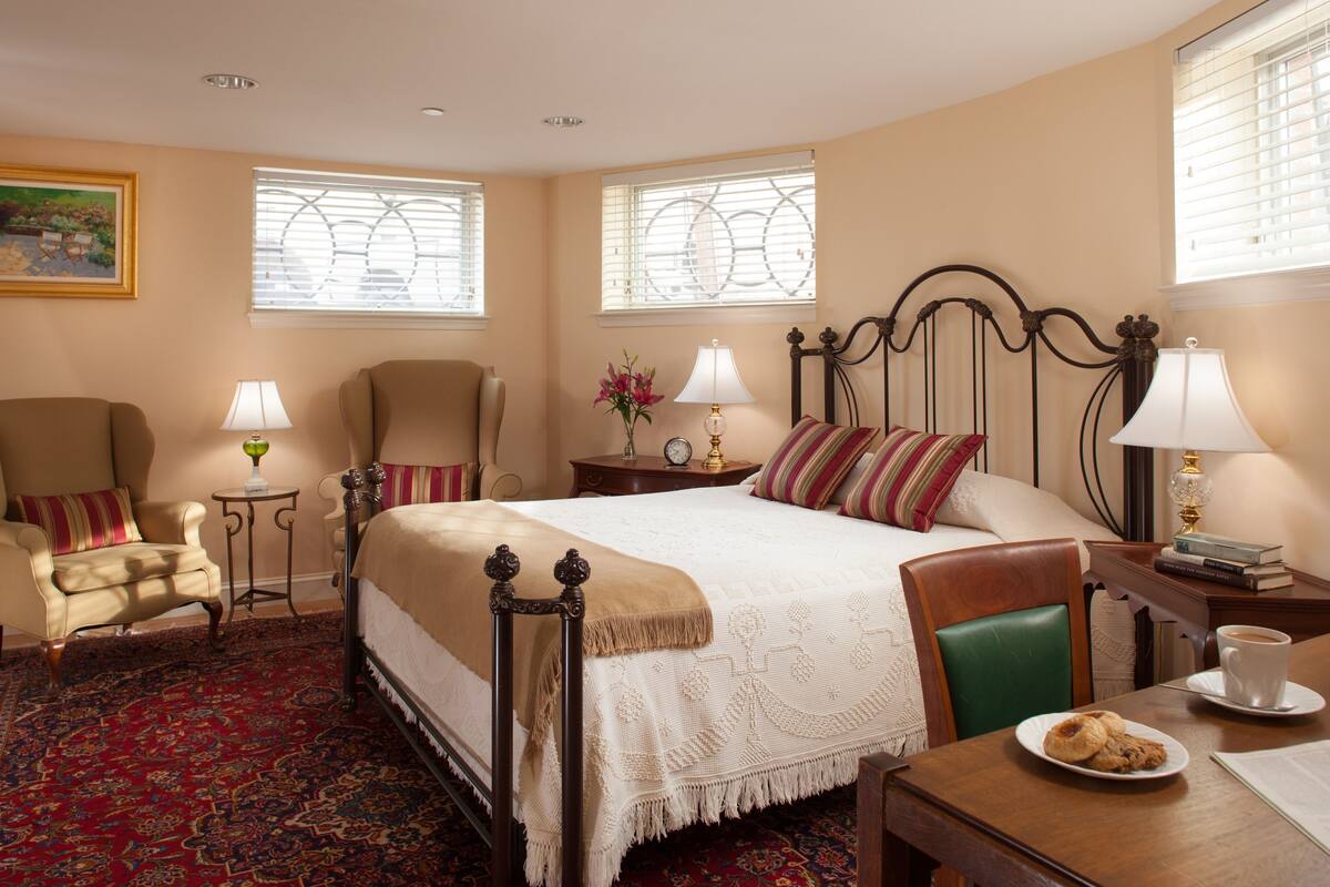 A hotel room with a large bed, two armchairs, a small table with a lamp, a side table with a lamp, a coffee table with a plate of cookies, and a window with blinds.