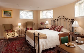 A hotel room with a large bed, two armchairs, a small table with a lamp, a side table with a lamp, a coffee table with a plate of cookies, and a window with blinds. - Woodley Park Guest House (Washington)