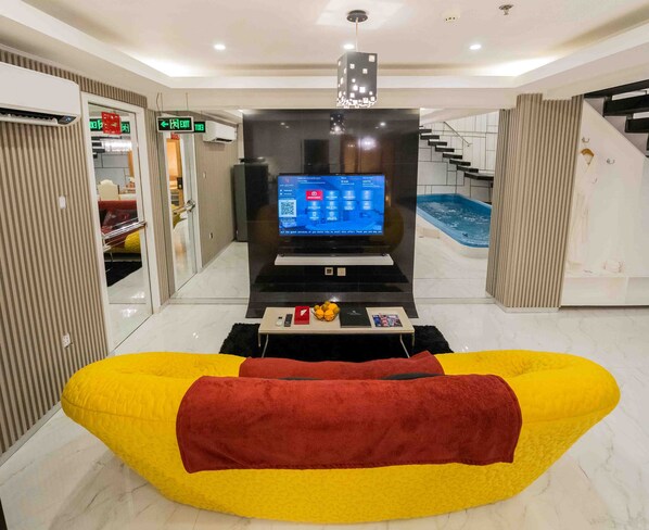 A hotel lobby with a flat-screen TV displaying a welcome message, a reception desk, a staircase, and a pool area.