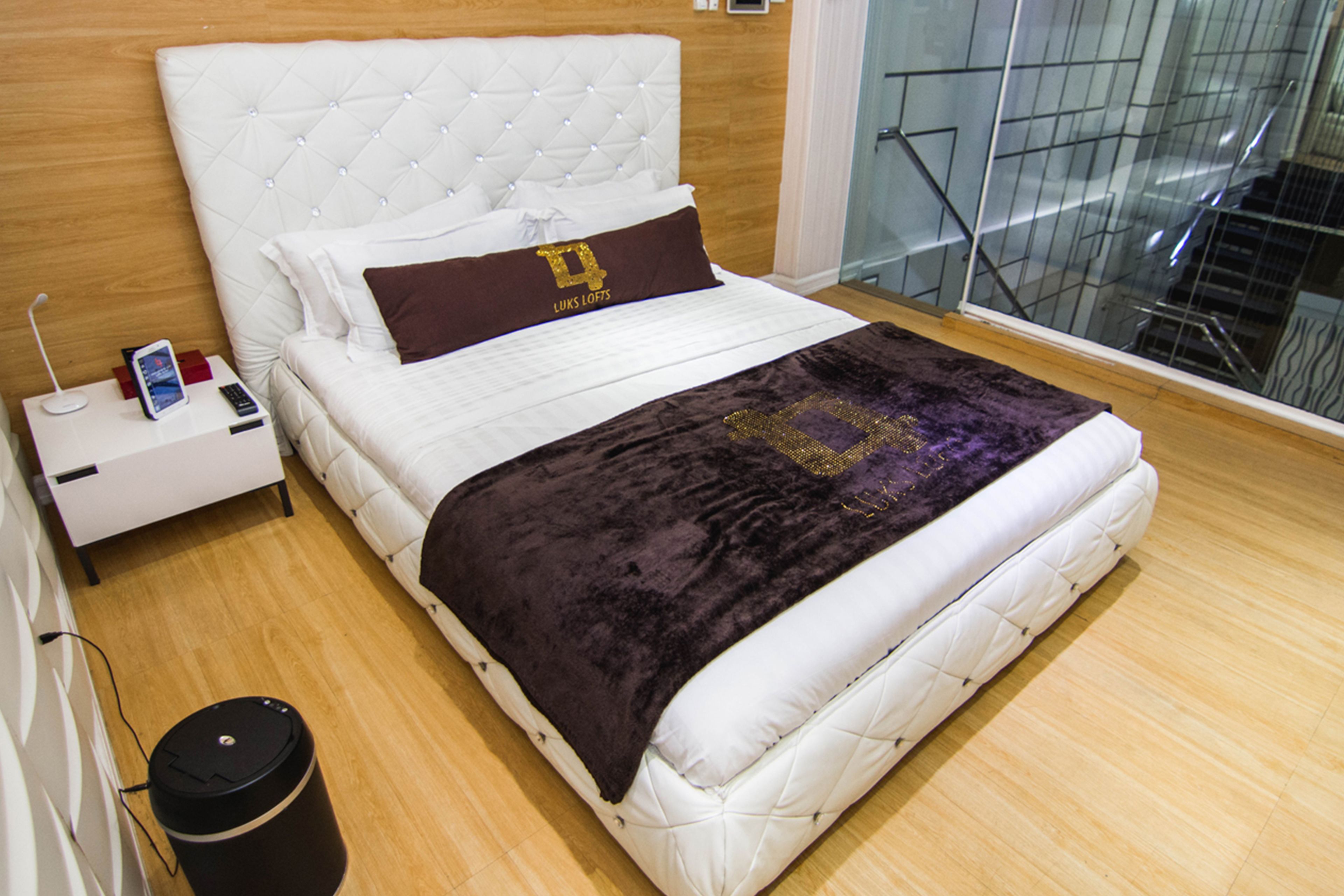 A neatly made bed with a white headboard and a dark purple blanket, a bedside table with a phone and a remote, and a trash bin.