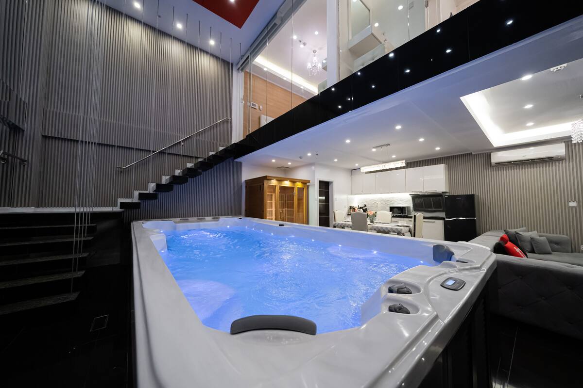Une piscine intérieure moderne, avec un escalier menant à un deuxième étage, un coin cuisine et un espace détente avec un canapé.