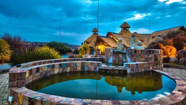 A pool with a stone border, a stone fountain, and a building with multiple roofs in the background.