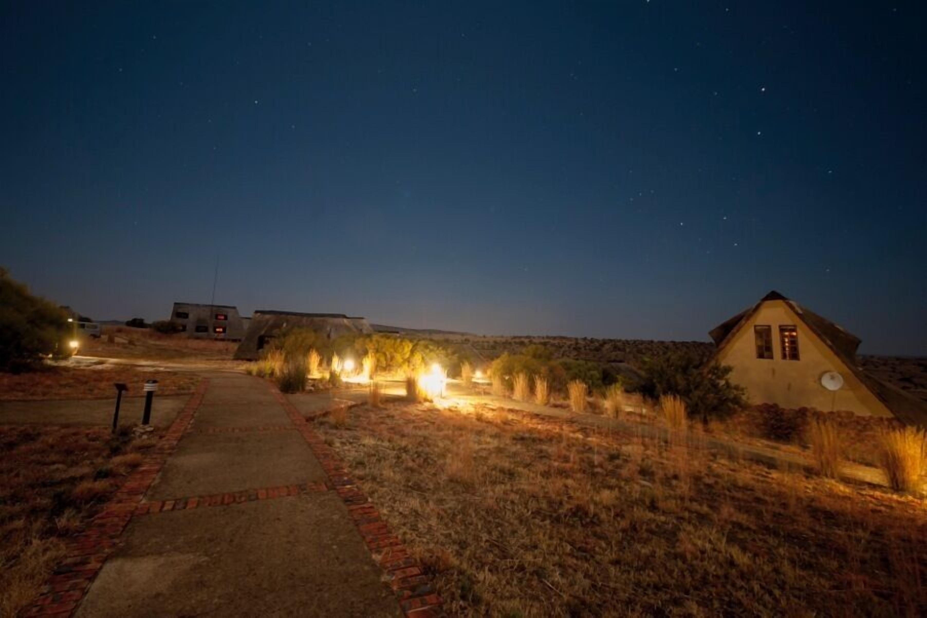 A pathway leading to a building with a triangular roof, surrounded by dry grass and illuminated by ground lights.