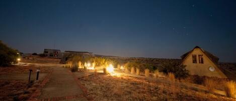 A pathway leading to a building with a triangular roof, surrounded by dry grass and illuminated by ground lights.
