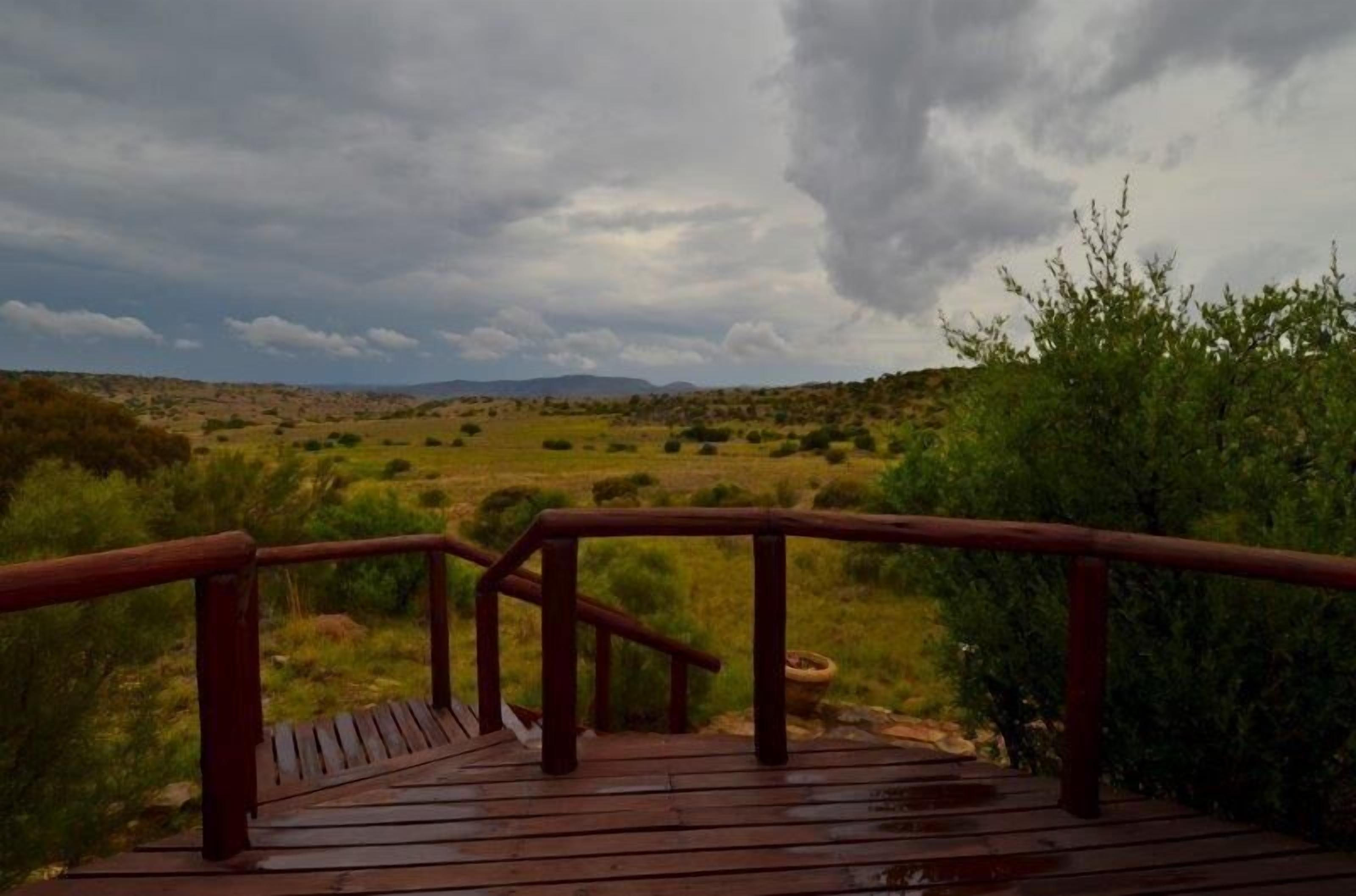 A wooden deck with a railing overlooking a grassy landscape with bushes and a distant mountain range under a cloudy sky.