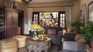 A cozy living room with a sofa, a coffee table with a vase of flowers, a bookshelf, and a large window with curtains.