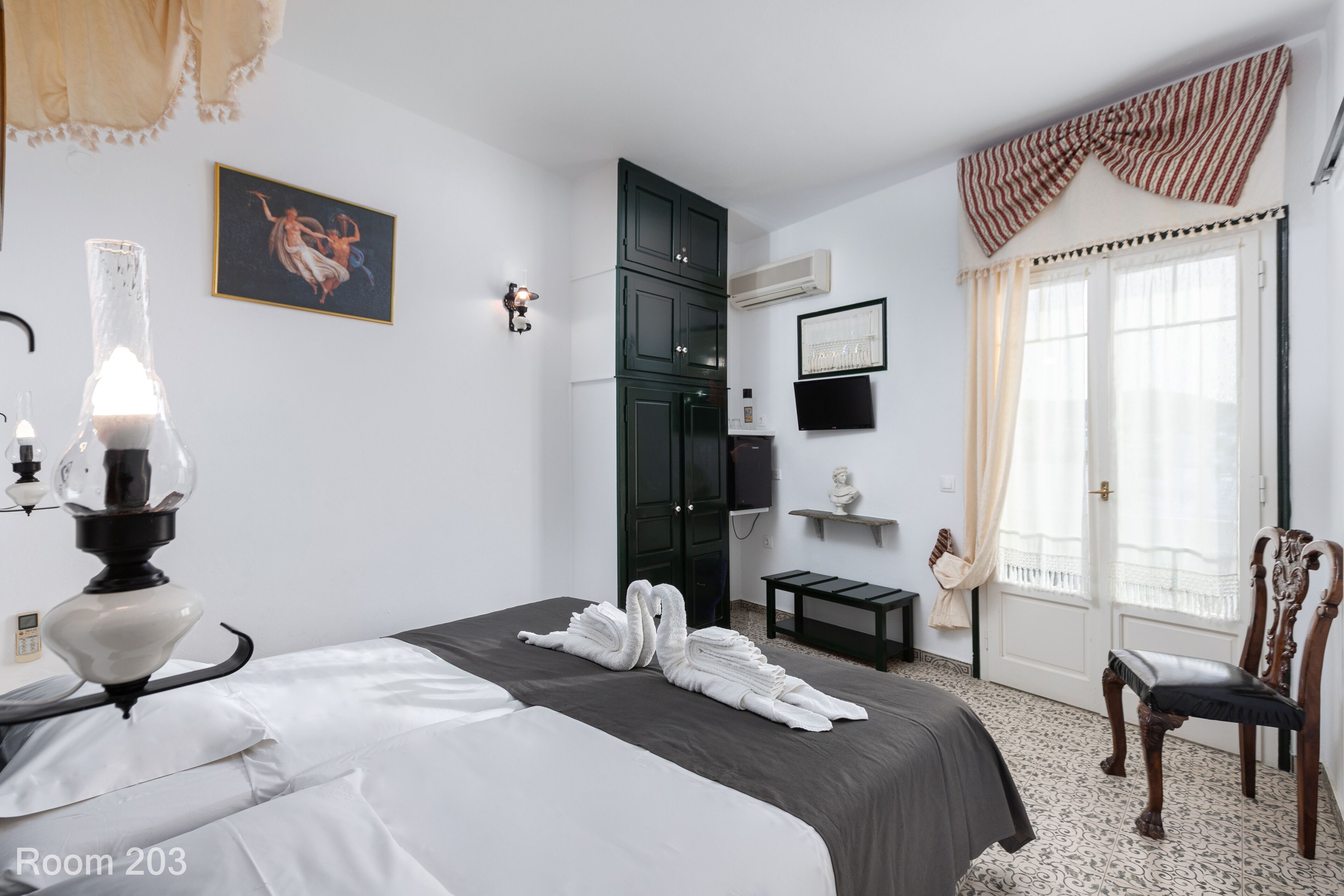 A hotel room with a bed, a desk, a chair, a TV, a wardrobe, a painting, and a window with curtains.