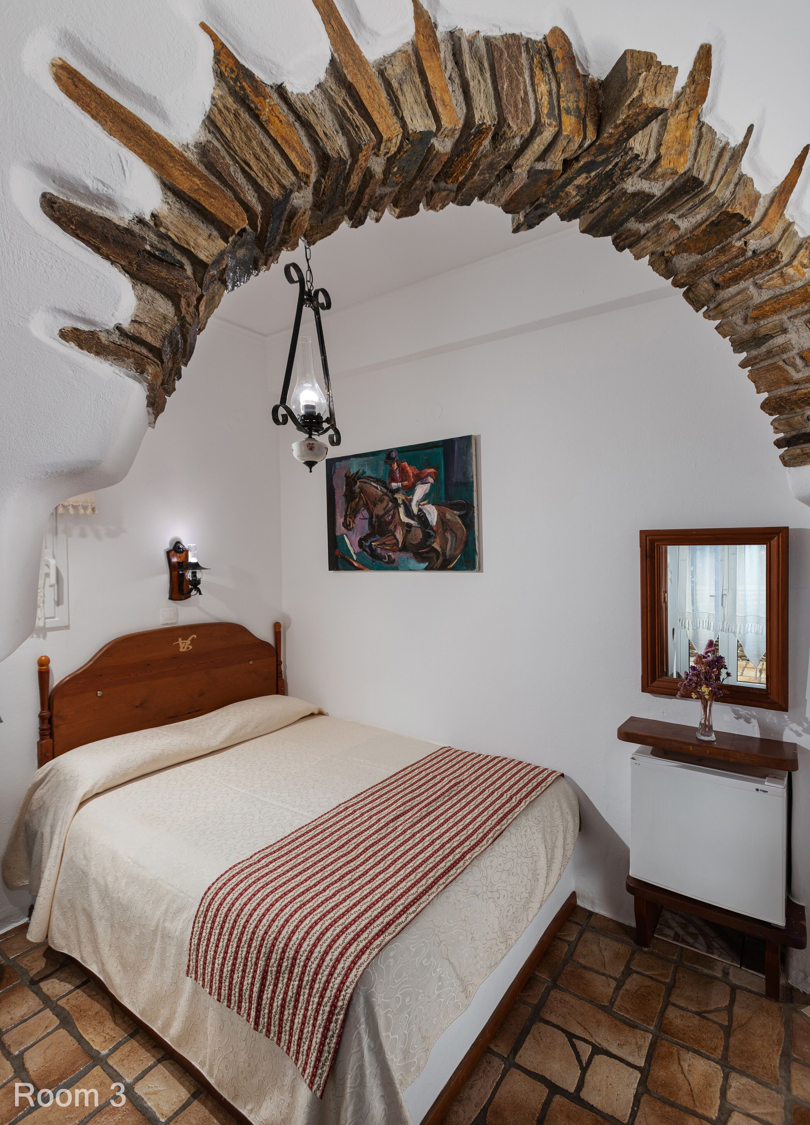 Un dormitorio con techo abovedado de piedra, una cama de madera, un cuadro, una lámpara de noche, un espejo y una mesita con un jarrón.