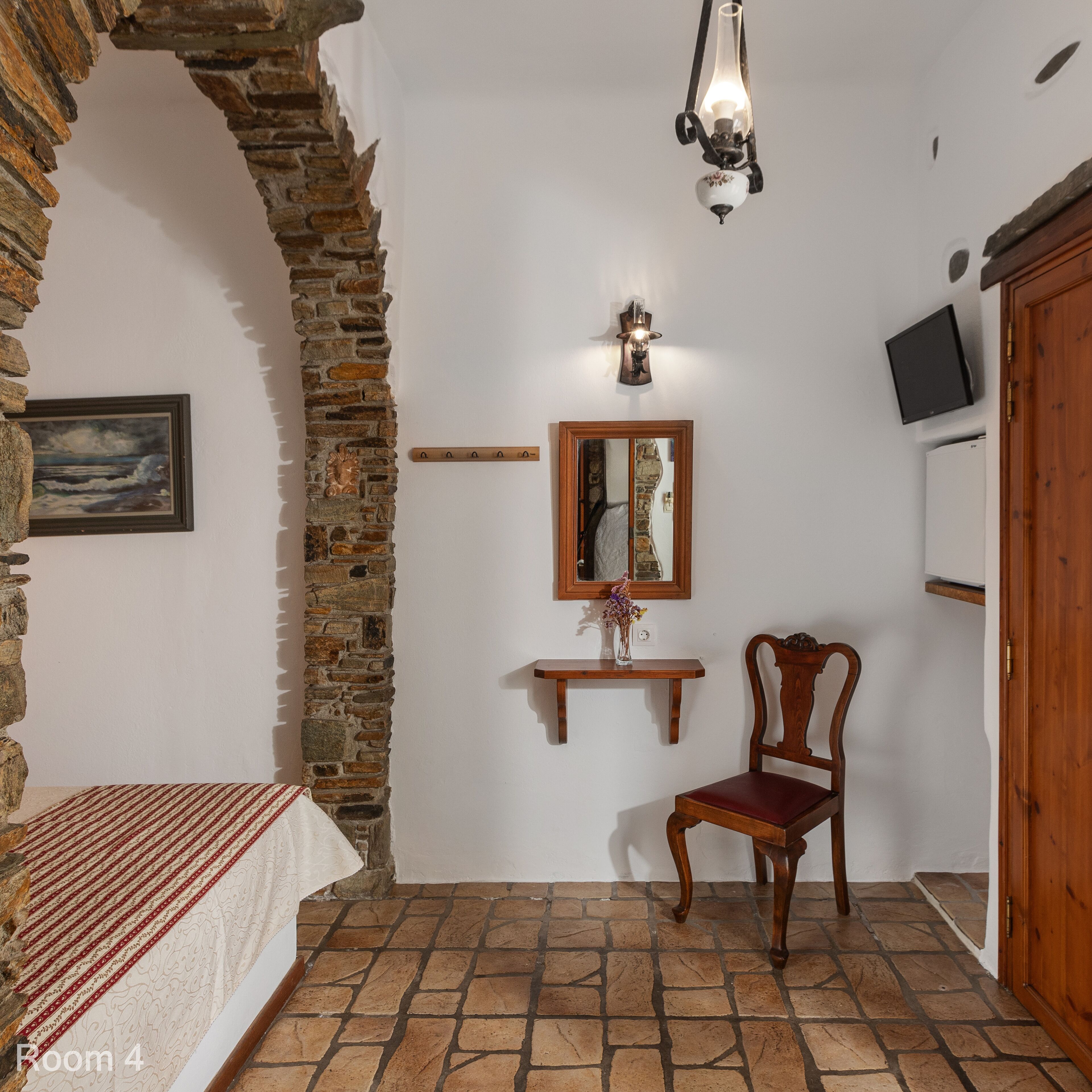 A room with a stone wall, a bed with a striped bedspread, a wooden chair, a small table with a vase, a wall-mounted light fixture, a mirror, and a television on the wall.
