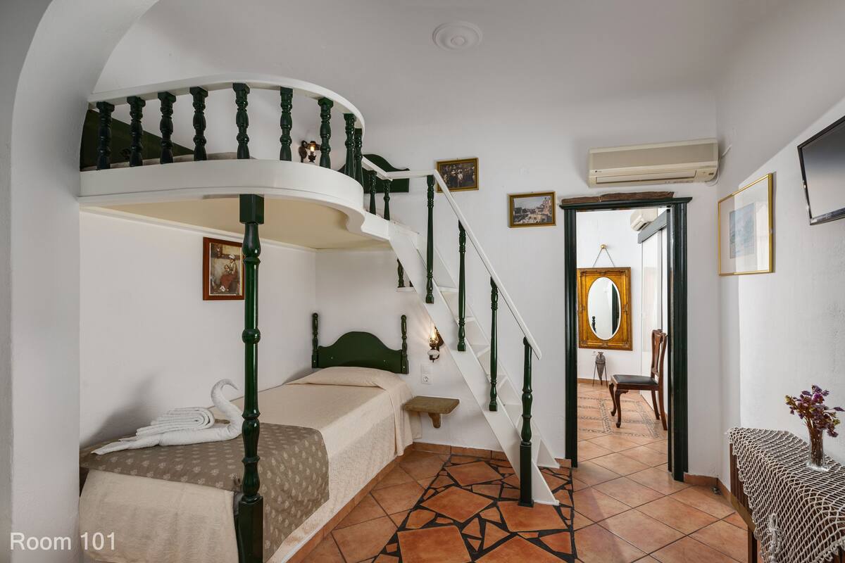 A room with a curved staircase, a bed with a green headboard, a small table with a vase, and a mirror on the wall.