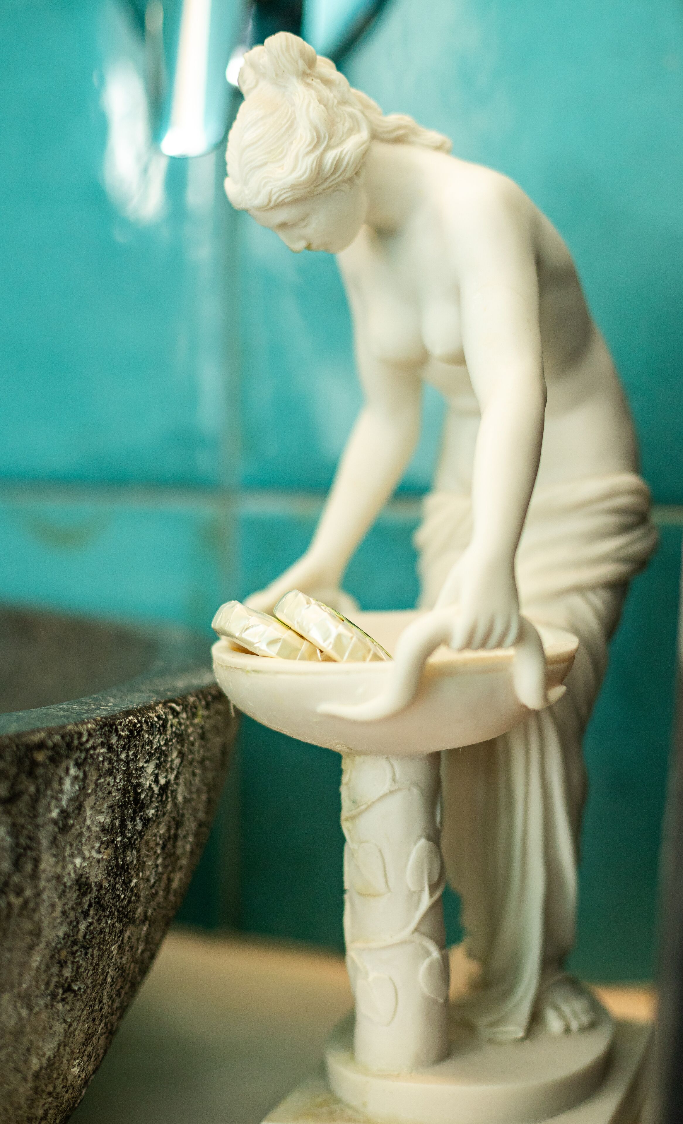 A white marble statue of a woman leaning over a basin, with a stone pedestal and a greenish-blue background.