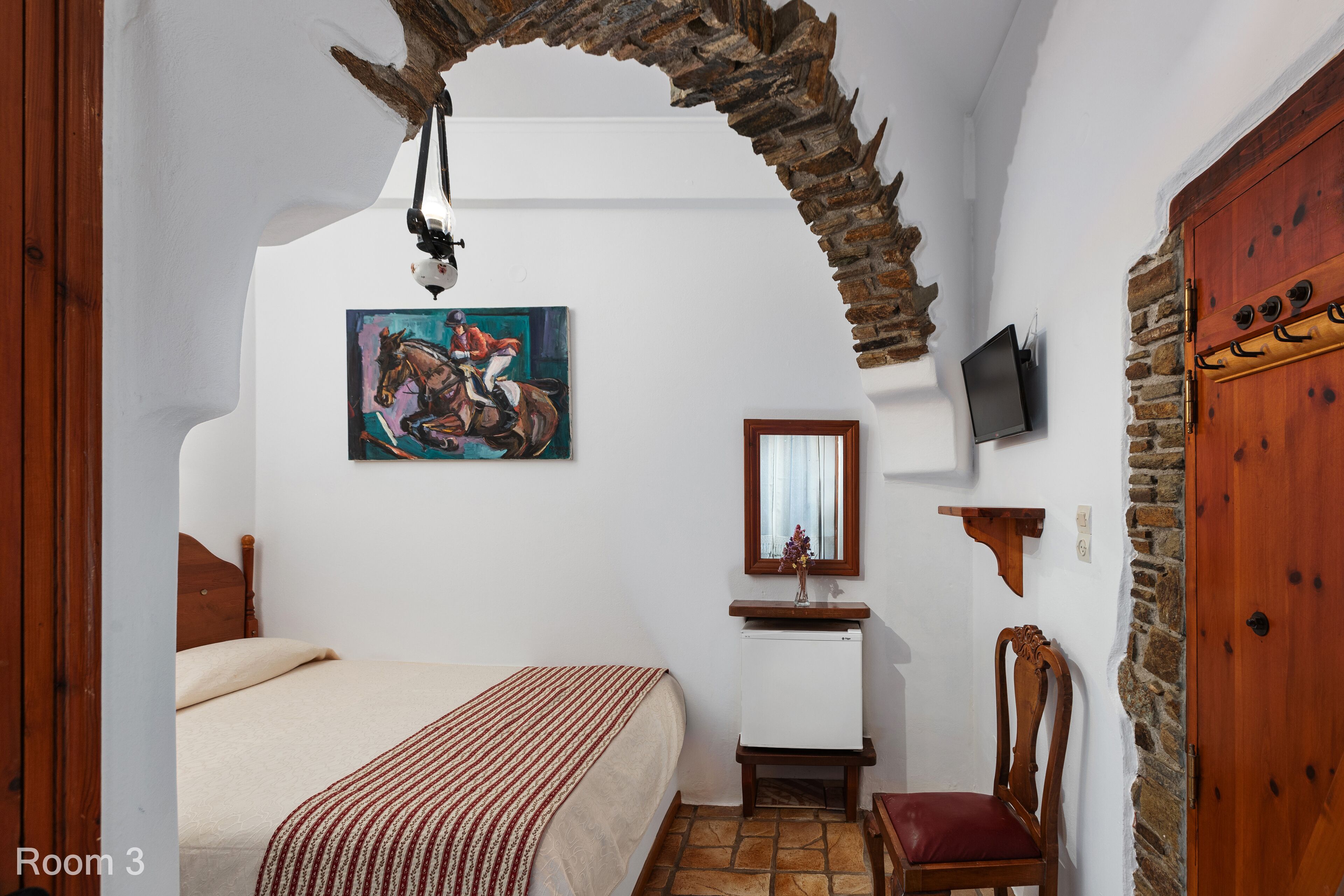 Un dormitori amb un llit, una cadira de fusta, una televisió, un quadre i una paret de pedra.