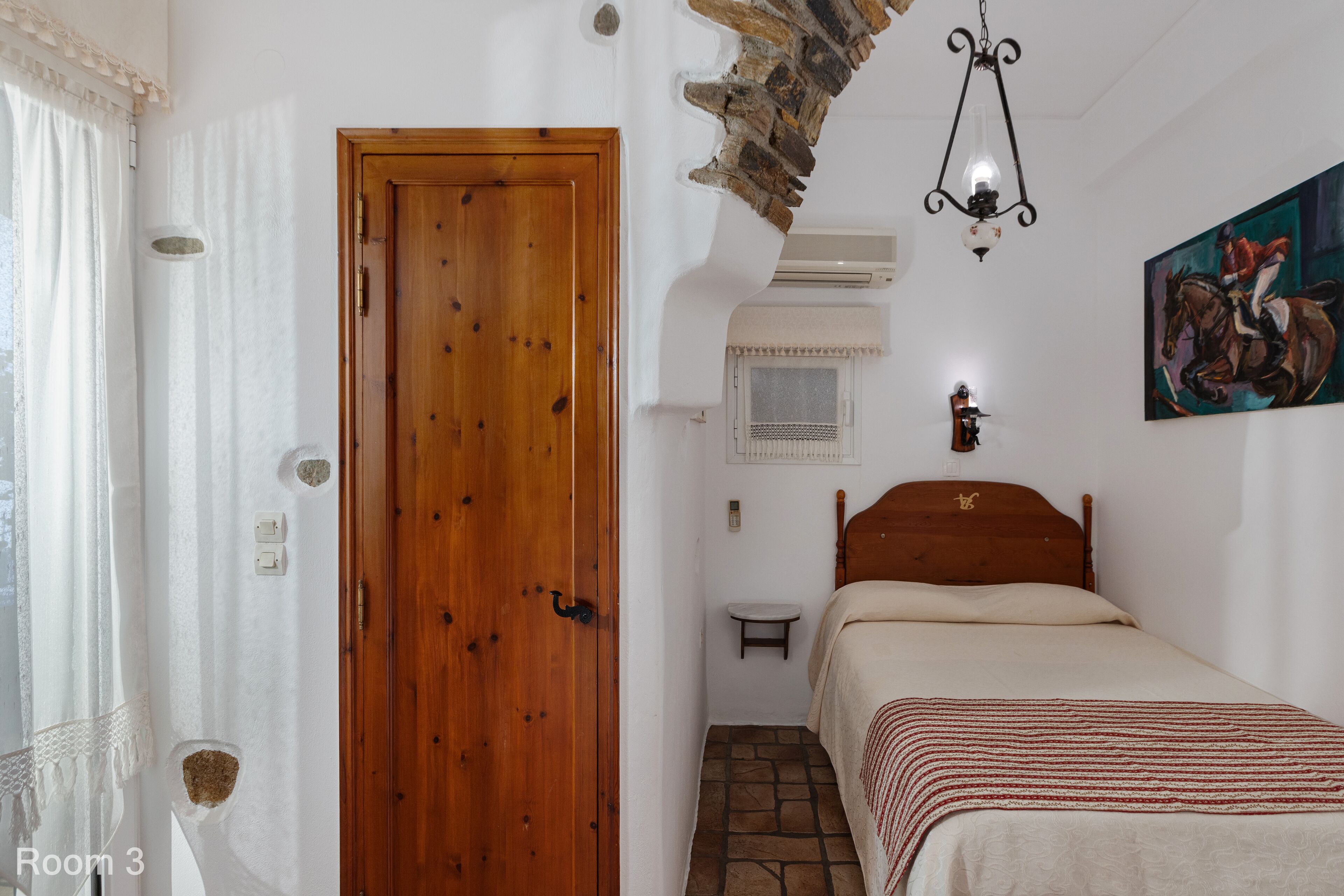 Un dormitorio con puerta de madera, una cama con manta a rayas, una mesita de noche pequeña, una lámpara de pared y un cuadro de un caballo.